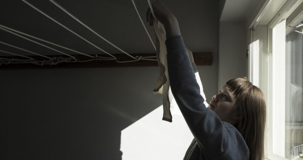 A person hangs laundry to dry in a drying room. The image shows ceiling-mounted clotheslines and daylight coming in through a window.