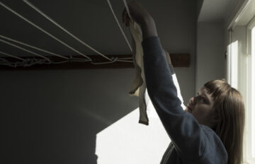 A person hangs laundry to dry in a drying room. The image shows ceiling-mounted clotheslines and daylight coming in through a window.