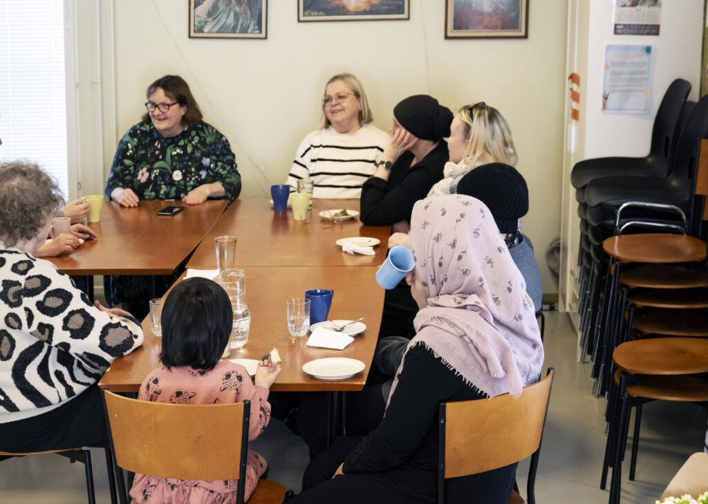 Asukastoimikunnan jäseniä istuu pöydän ääressä. Pöydällä on kuppeja ja lautasia.