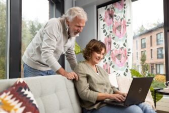A person sits on a sofa using a laptop, while another person stands beside them looking at the screen.