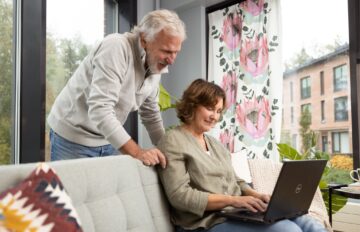 En person sitter i en soffa och använder en bärbar dator, en annan person står bredvid och tittar på skärmen.