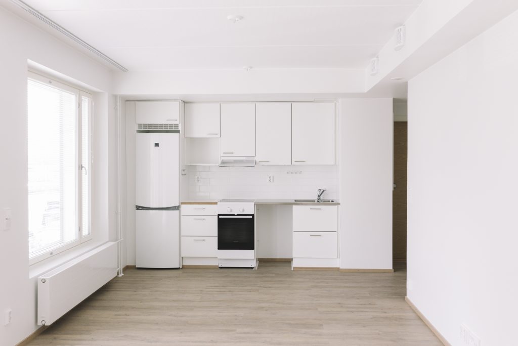 Huonenäkymä, jossa valkoisten seinien keskellä valkoinen keittiöseinäke ja vaalea lattia.