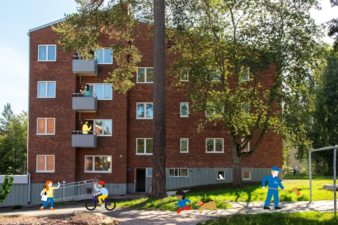 Punatiilinen kerrostalo puiden keskellä kesäisessä pihassa. Parvekkeilla seisoo asukkaita, ja pihan käytävällä kulkee ihmisiä: yksi pyörällä, yksi ulkoiluttaa koiraa ja yksi lakaisee käytävää.