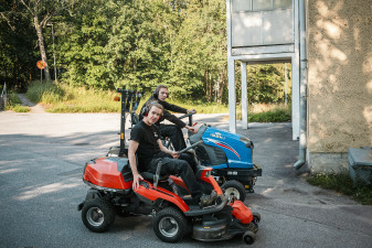Kaksi huollon kesätyötekijää ajamassa päältäajettavia ruohonleikkureita kerrostalon pihalla kesäisessä ympäristössä.