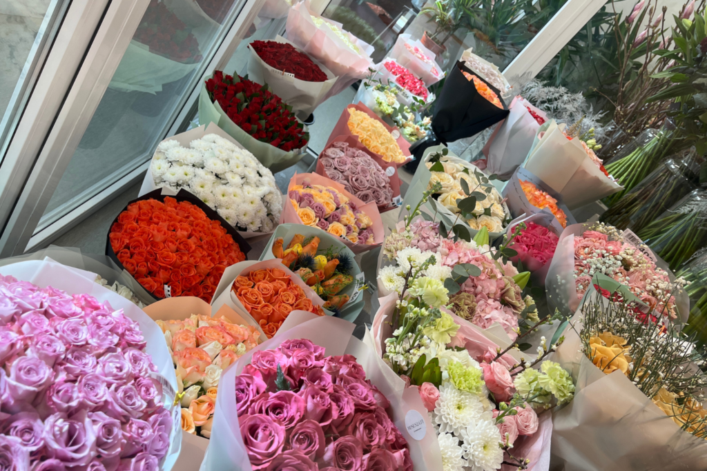 Värikkäitä kukkakimppuja kukkakaupan kylmiössä, mukana ruusuja, neilikoita ja muita leikkokukkia eri sävyissä.