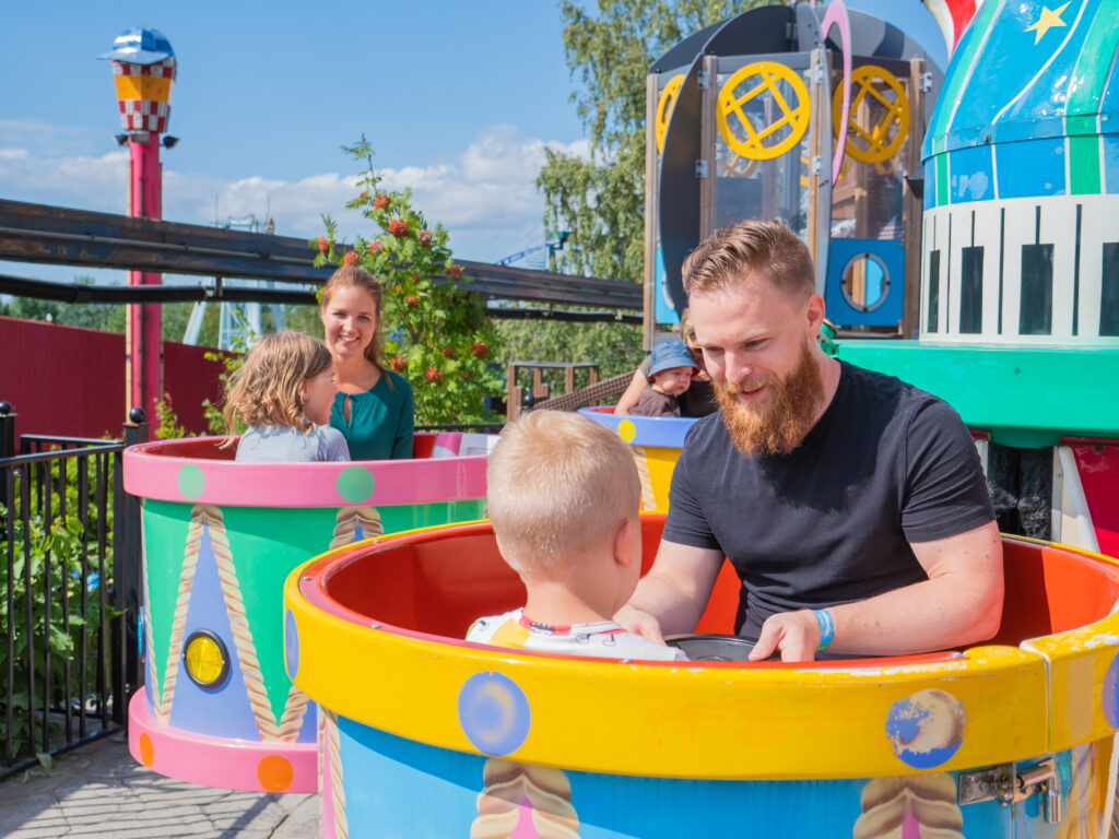Aikuisia ja lapsia värikkäässä huvipuistolaitteessa, jossa istutaan pyörivissä kupeissa. Etualalla aikuinen ja pieni lapsi istuvat keltaisessa ja sinisessä kupissa, taustalla muita kuppeja ja huvipuistolaitteita aurinkoisena päivänä.