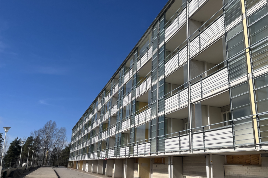 Pitkä, vaalea kerrostalo, jossa on useita lasitettuja parvekkeita. Rakennus kuvattu alhaalta kadun suunnasta kirkkaana ja aurinkoisena päivänä, sininen taivas taustalla.