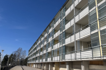 Pitkä, vaalea kerrostalo, jossa on useita lasitettuja parvekkeita. Rakennus kuvattu alhaalta kadun suunnasta kirkkaana ja aurinkoisena päivänä, sininen taivas taustalla.