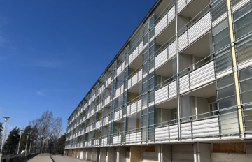 Pitkä, vaalea kerrostalo, jossa on useita lasitettuja parvekkeita. Rakennus kuvattu alhaalta kadun suunnasta kirkkaana ja aurinkoisena päivänä, sininen taivas taustalla.