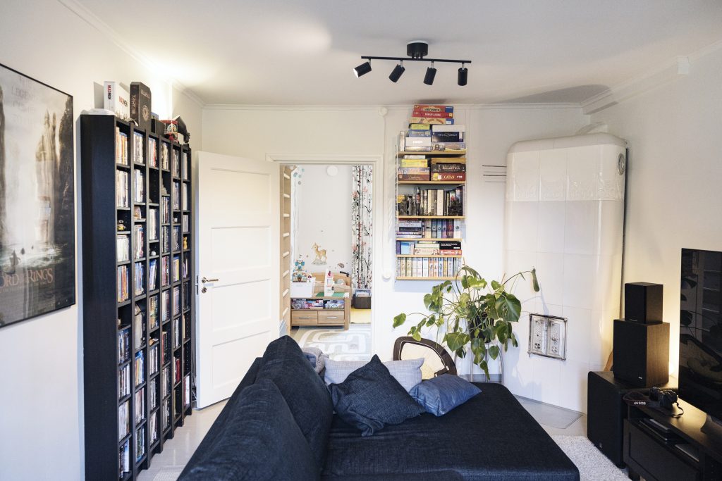 A linving room with a sofa, tiled stove, television and bookshelves.