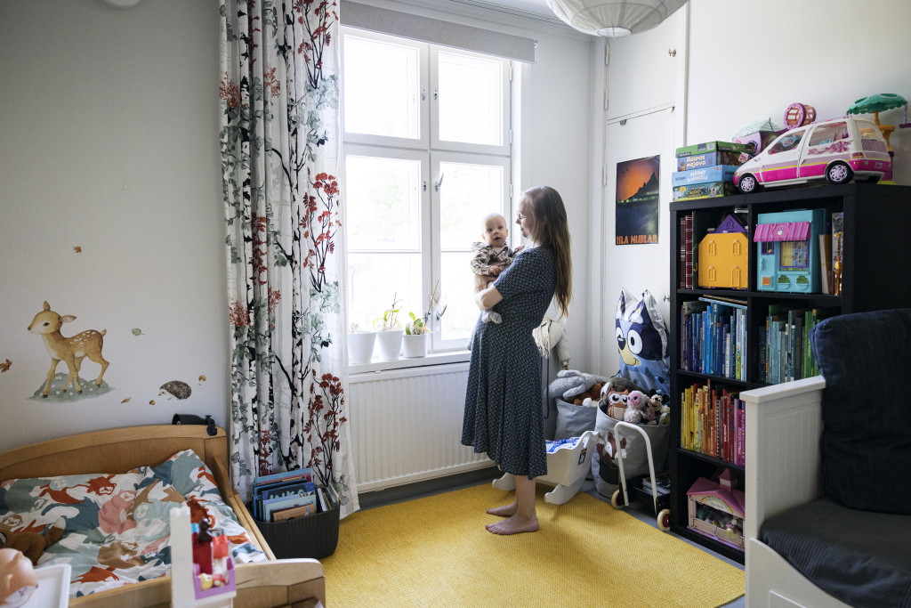 Nainen, jolla on vauva sylissään, seisoo huoneessa ikkunan edessä. Lasten sänky, kirjahylly ja muita lastentavaroita.
