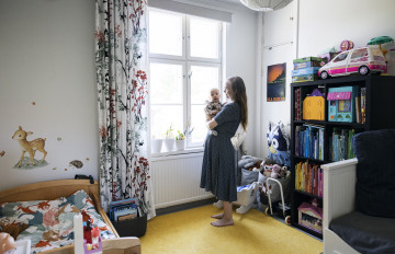 Nainen, jolla on vauva sylissään, seisoo huoneessa ikkunan edessä. Lasten sänky, kirjahylly ja muita lastentavaroita.