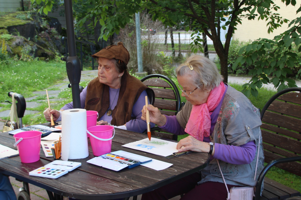 Kaksi ikäihmistä maalaa paperille vesiväreillä puutarhassa pöydän ääressä.