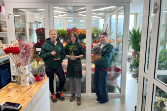 Kolme kukkakaupan työntekijää seisoo kylmiön ovien edessä kukkakaupassa. Jokainen pitää käsissään ruukkukasvia. Taustalla näkyy runsaasti värikkäitä leikko- ja viherkasveja sekä kukkakaupan myyntitilaa.