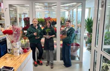 Kolme kukkakaupan työntekijää seisoo kylmiön ovien edessä kukkakaupassa. Jokainen pitää käsissään ruukkukasvia. Taustalla näkyy runsaasti värikkäitä leikko- ja viherkasveja sekä kukkakaupan myyntitilaa.