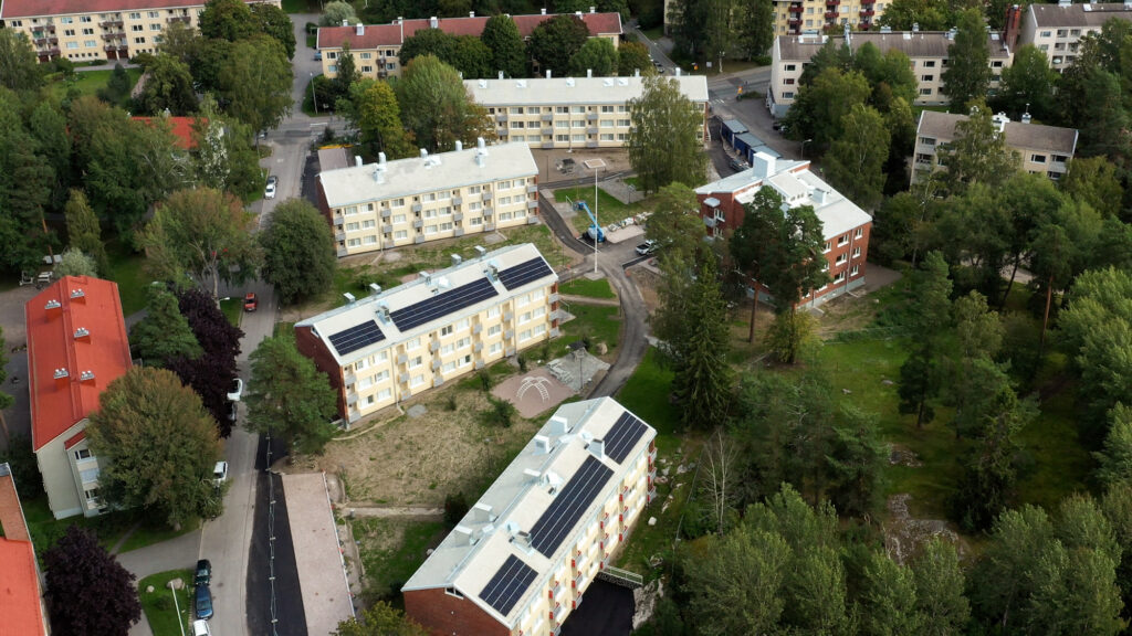 Ilmakuva Maunulan Heka-kerrostaloista: vaaleita 3–4-kerroksisia asuinrakennuksia, joiden katoilla on aurinkopaneeleja. Rakennuksia ympäröivät puut, pihatiet ja oleskelualueet, ja korttelin keskellä näkyy pieni leikkipaikka sekä rakennustyökone.