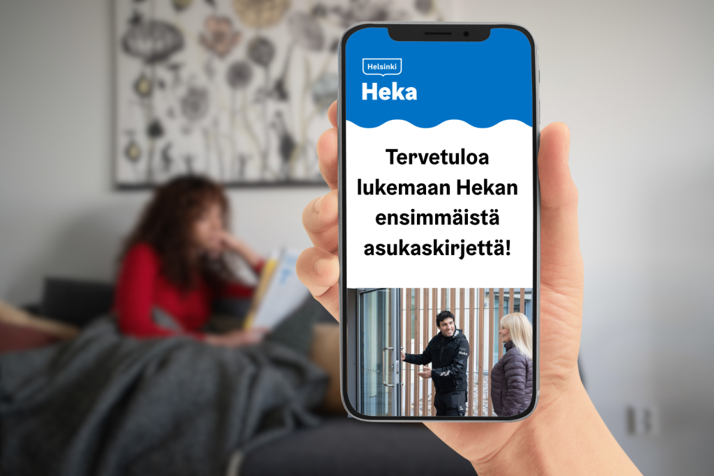 Kädessä puhelin, jonka näytöllä teksti: Tervetuloa lukemaan Hekan ensimmäistä asukaskirjettä. Taustalla nainen istuu sohvalla.