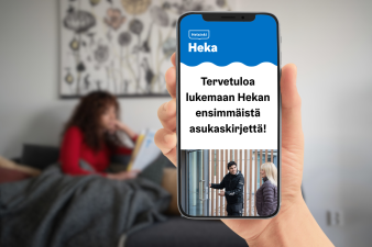 Kädessä puhelin, jonka näytöllä teksti: Tervetuloa lukemaan Hekan ensimmäistä asukaskirjettä. Taustalla nainen istuu sohvalla.