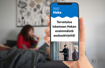 Kädessä puhelin, jonka näytöllä teksti: Tervetuloa lukemaan Hekan ensimmäistä asukaskirjettä. Taustalla nainen istuu sohvalla.