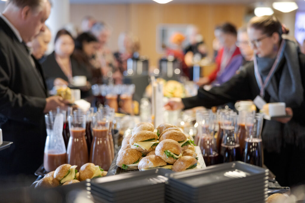 Aamiaistarjoilu: pöydällä sämpylöitä ja juomia, osallistujat ottavat tarjottavaa.