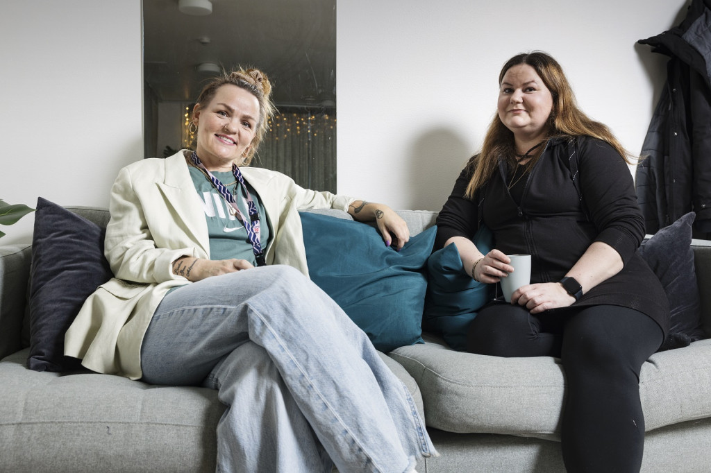 Neighbours Linda Granström and Riina Puotsaari are sitting together on a grey couch. Granström has light-coloured clothes and her hair tied up in a bun. Puotsaari has black clothes and long, brown hair. Both are holding a coffee cup.