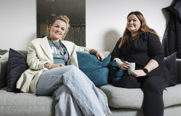 Neighbours Linda Granström and Riina Puotsaari are sitting together on a grey couch. Granström has light-coloured clothes and her hair tied up in a bun. Puotsaari has black clothes and long, brown hair. Both are holding a coffee cup.
