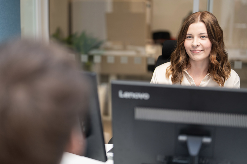 Nuori henkilö istuu tietokoneen ääressä ja katsoo hymyillen vastapäätä istuvaan henkilöä.