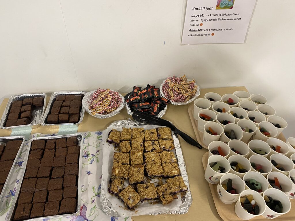 Juhlapöydälle on katettu brownies-leivonnaisia, marjapiirakkaa, makeisia ja karkkikuppeja osallistujille. Seinällä on ohjeistus karkkikuppien jakamista varten.