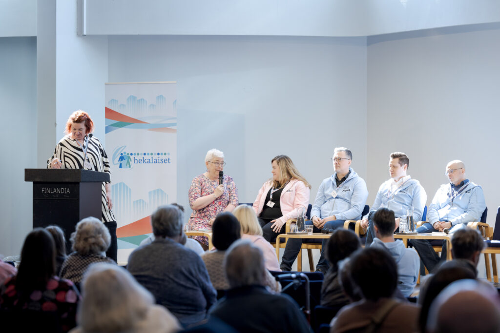 Paneelikeskustelu käynnissä lavalla, edessä yleisöä ja taustalla tapahtuman banderolli.