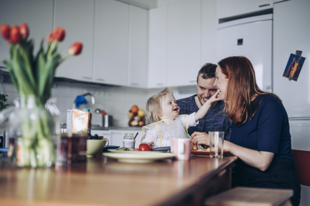 Lapsiperhe viettämässä aikaa ruokapöydän ääressä. Pöydällä on aamiaistarvikkeita ja kukkaruukussa tulppaaneita.