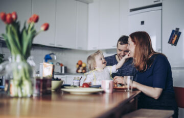 Lapsiperhe viettämässä aikaa ruokapöydän ääressä. Pöydällä on aamiaistarvikkeita ja kukkaruukussa tulppaaneita.