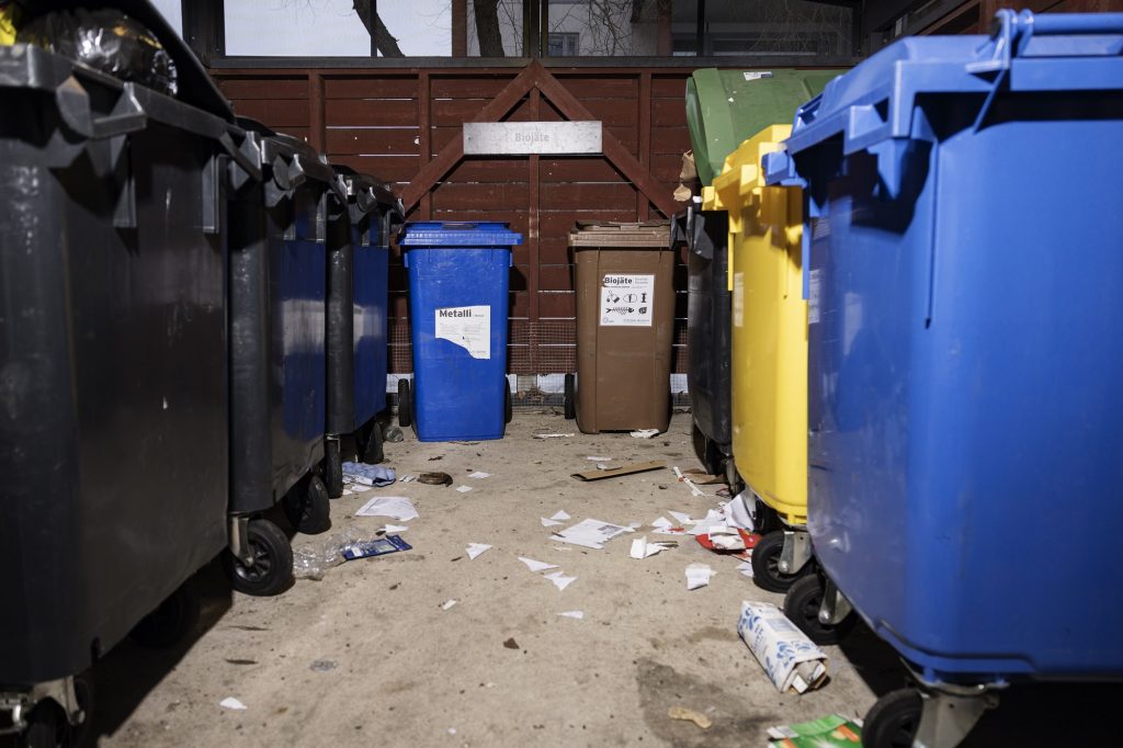 Det finns skräp på avfallsutrymmets golv. På bilden finns svarta insamlingskärl för blandavfall, ett brunt insamlingskärl för bioavfall, ett gult insamlingskärl för plast och ett blå insamlingskärl för metall. 