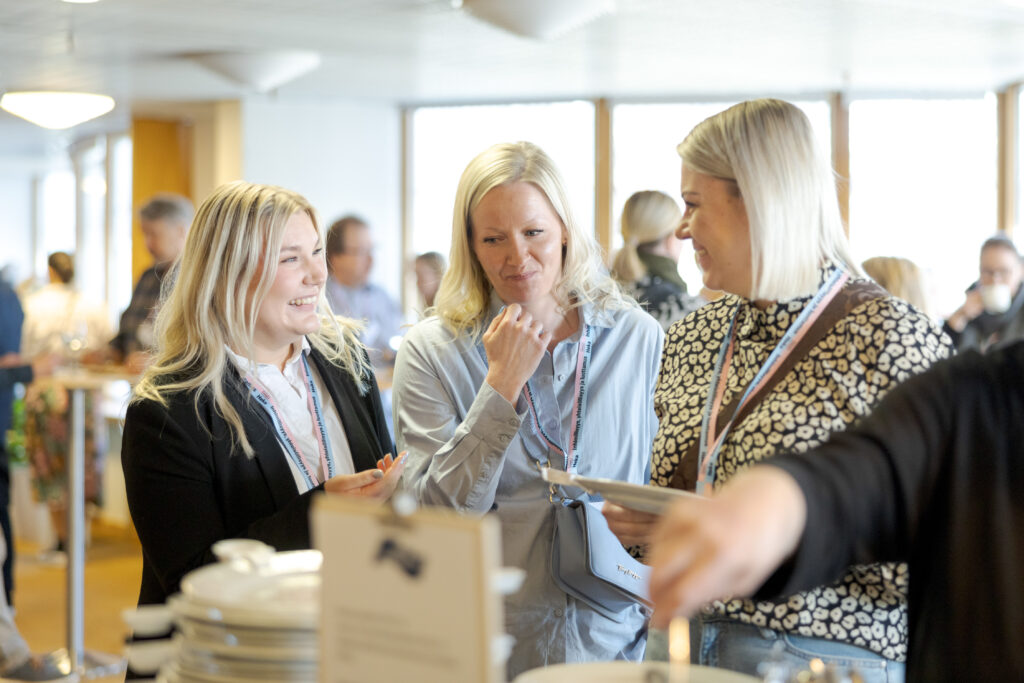 Kolme tapahtumaosallistujaa keskustelee seisten tarjoilupöydän äärellä sisätilassa. Heillä on kaulanauhat nimikorteilla, ja taustalla näkyy muita osallistujia epäselvästi.