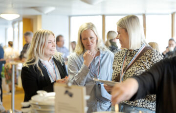 Kolme tapahtumaosallistujaa keskustelee seisten tarjoilupöydän äärellä sisätilassa. Heillä on kaulanauhat nimikorteilla, ja taustalla näkyy muita osallistujia epäselvästi.