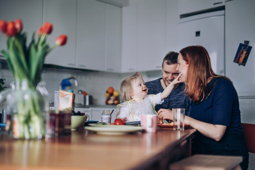 A small child wearing a bib sits at a kitchen table and reaches toward her mom seated nearby. The table is set with plates, cups, food and drinks. Dad sits behind the child. In the foreground, a vase with tulips is slightly out of focus.