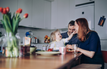 A small child wearing a bib sits at a kitchen table and reaches toward her mom seated nearby. The table is set with plates, cups, food and drinks. Dad sits behind the child. In the foreground, a vase with tulips is slightly out of focus.