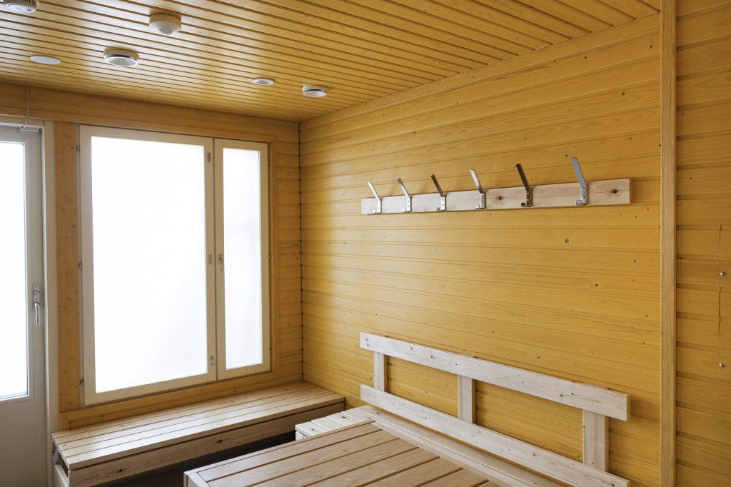 Kuvassa on saunan pukuhuone, jonka seinät on maalattu keltaiseksi. Huoneessa on iso ikkuna.