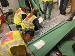 Huomioliiveihin pukeutuneet osallistujat rakentavat kerrossänkyjä väestönsuojassa.