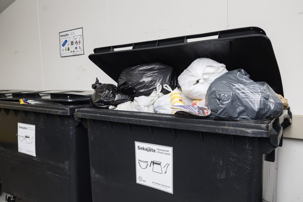 The lid of a black mixed waste container cannot be closed because the container is too full of trash bags. There is another waste container next to it, the lid of which closes.