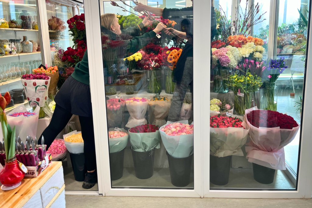 Kukkakaupan kylmiössä työntekijät ottavat leikkokukkia hyllystä. Etualalla ja taustalla näkyy runsaasti värikkäitä kukkia ja valmiita kimppuja.