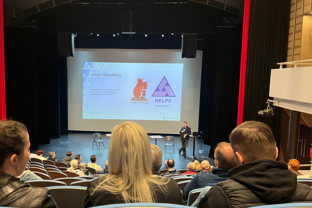 Yleisö seuraa turvallisuusaiheista esitystä auditoriossa. Lavalla esiintyjä puhuu suuren valkokankaan edessä.