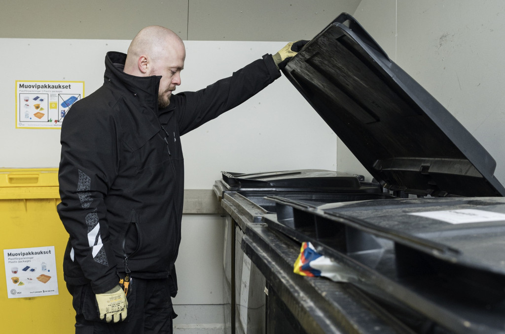 Maintenance Master Ville Laamanen is holding the lid of a black mixed waste container open with his left hand and looking inside the container. The background features more black waste containers and a yellow plastic waste container. The picture was taken at a Heka waste collection point.