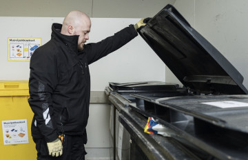 Underhållsmästare Ville Laamanen håller locket till ett svart insamlingskärl för blandavfall öppet med sin vänstra hand och tittar in i kärlet. I bakgrunden finns fler svarta avfallskärl och ett gult insamlingskärl för plast. Bilden har tagits vid en av Hekas avfallspunkter.