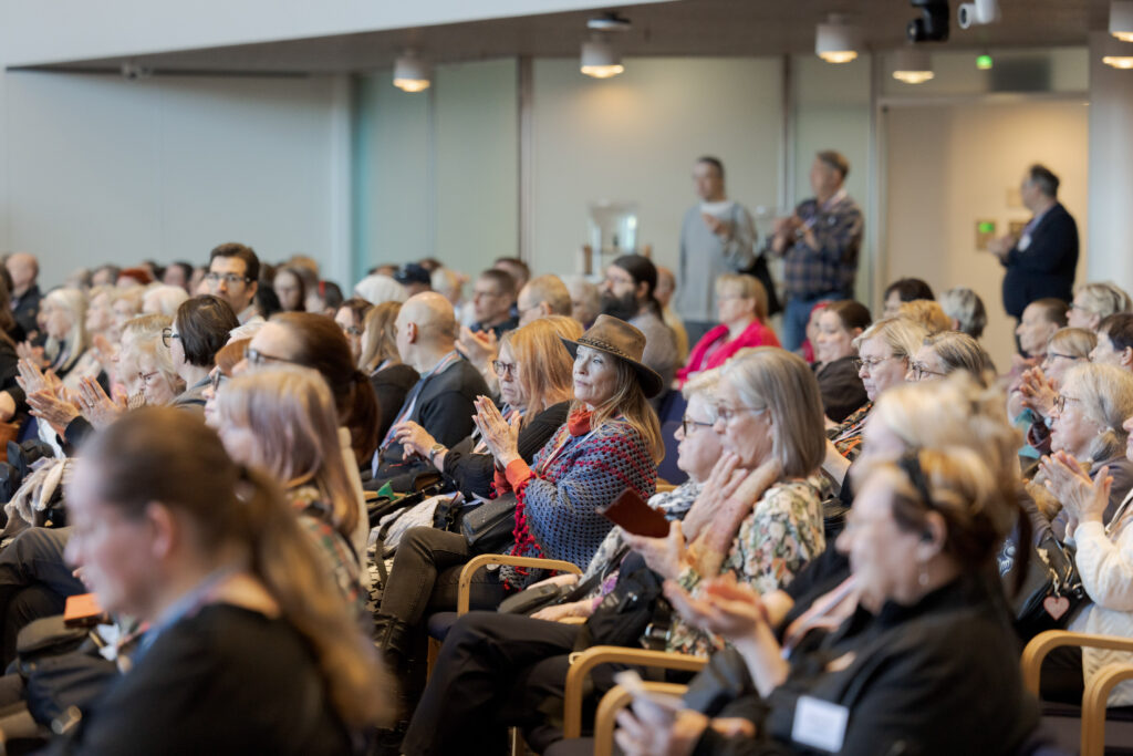 Yleisöä salissa katsomassa esitystä, osa taputtaa.